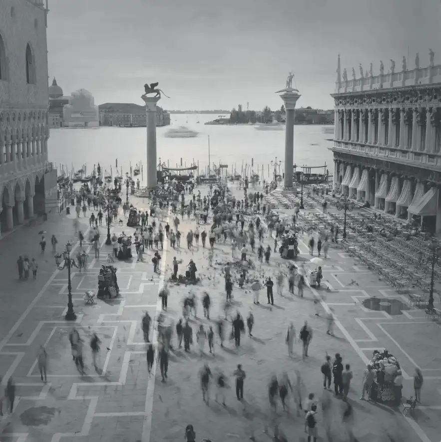 Long-exposure pedestrians cross Piazzetta San Marco between the columns and waterfront in Venice.