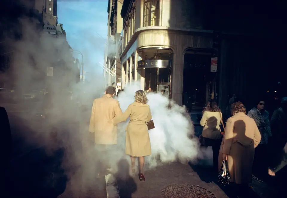 Two people in camel-colored coats walk into sunlit steam on a New York sidewalk.