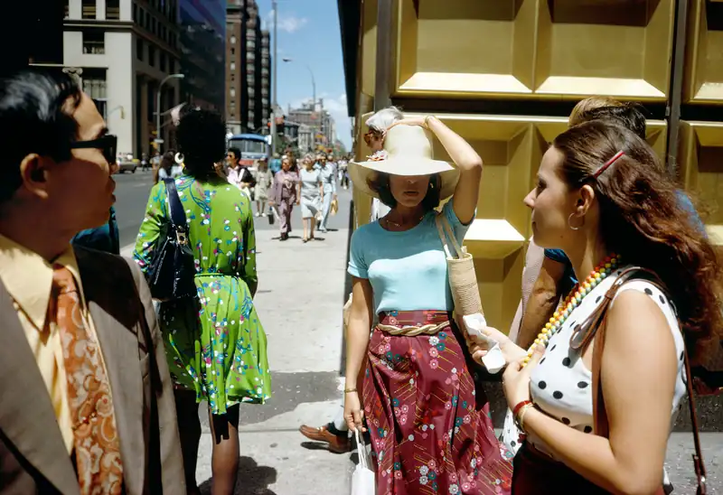 A woman holds her white hat at a busy New York corner beside a golden building facade.