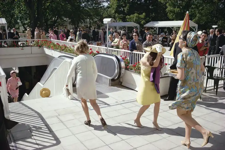Women in colorful summer dresses pass an outdoor escalator in a busy Paris street scene.