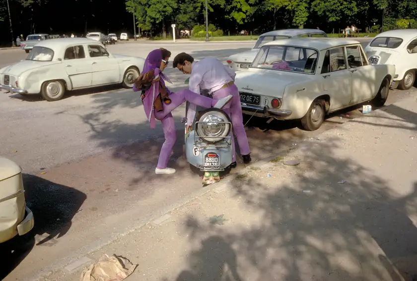 A couple dressed in purple stand over a scooter beside parked cars in Paris.