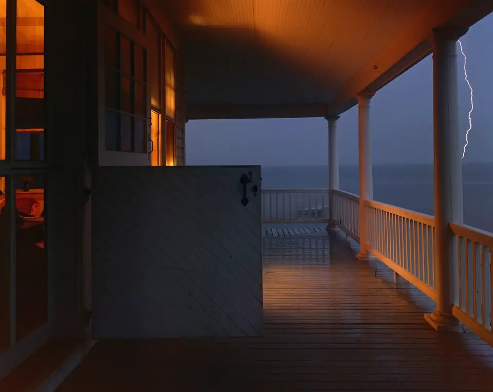 Warm light fills a wooden porch overlooking blue water while lightning crosses the sky.