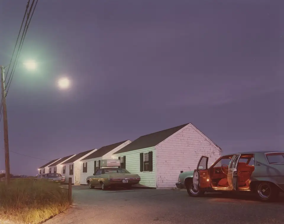A car with an open door and red interior sits beside white cottages under a violet evening sky.