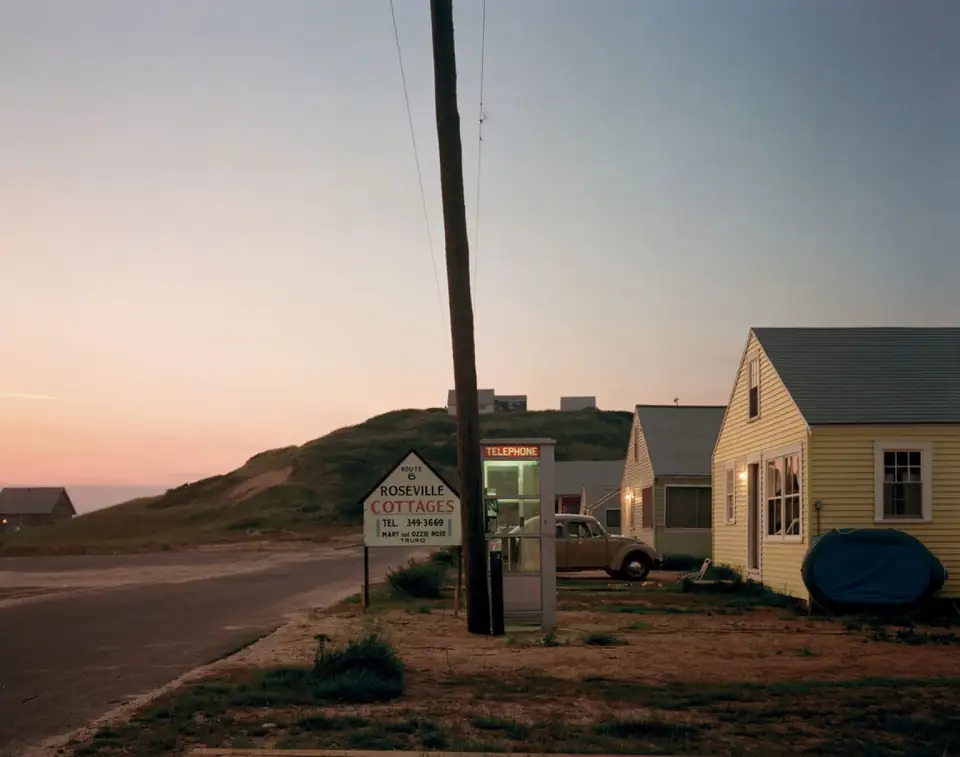 Small white roadside cottages and utility poles stand against a pale pink evening sky in Truro.
