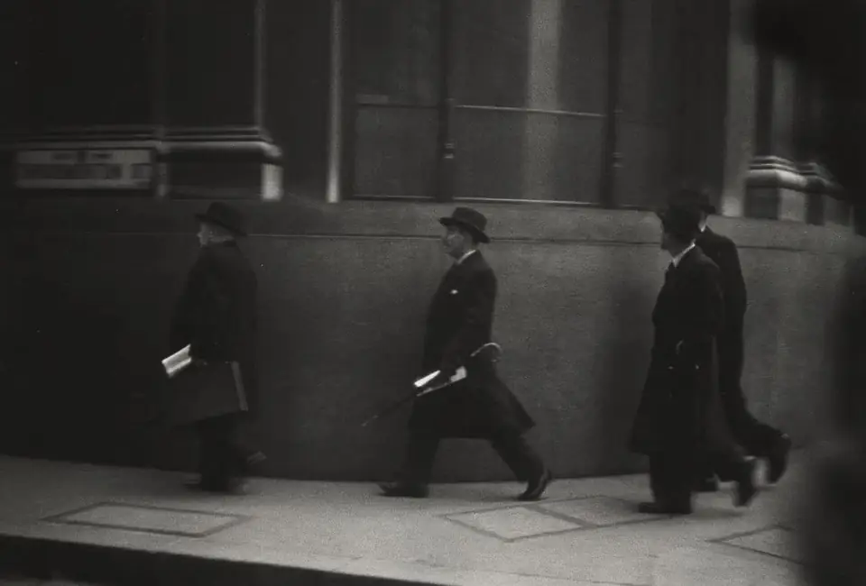 Three men in dark coats and hats walk along a London pavement beside a stone building.
