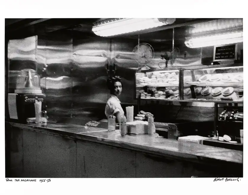 A woman stands behind a railway-station coffee counter amid blurred reflections and ceiling lights.