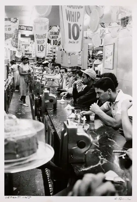 Customers sit along a drugstore lunch counter beneath crowded signs and ceiling lights.