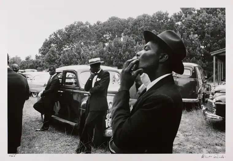 A man in a dark suit and hat stands in profile beside parked cars and a group of mourners.