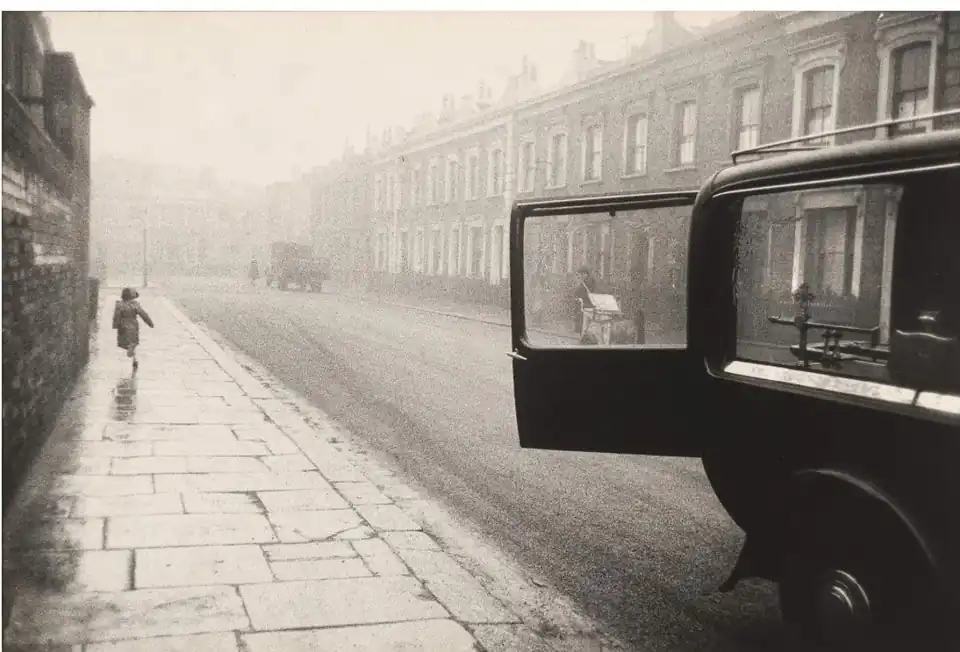 A child runs along a misty London pavement while the open door of a hearse frames a distant street worker.