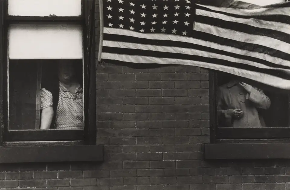 An American flag hangs across a brick building, partly hiding people standing in two windows.
