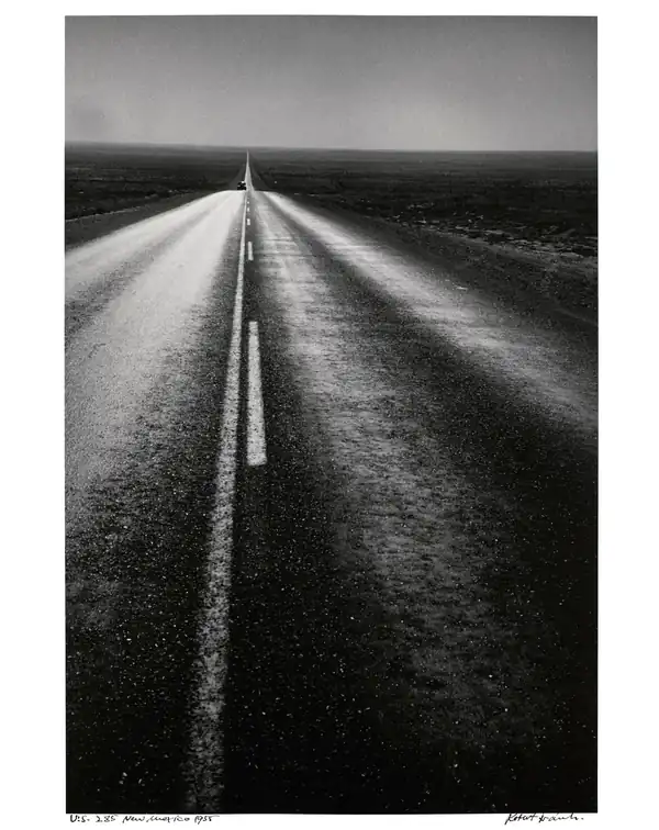A straight two-lane highway stretches through a dark, open New Mexico landscape to the horizon.