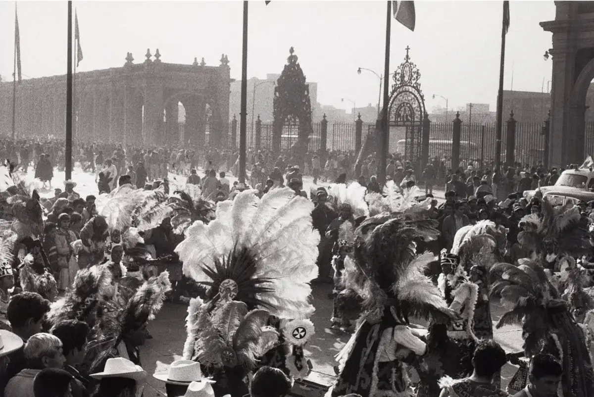 A large crowd gathered in front of an ornate gate with decorative ironwork, some people wearing traditional costumes and feathered headdresses in a street parade or festival.