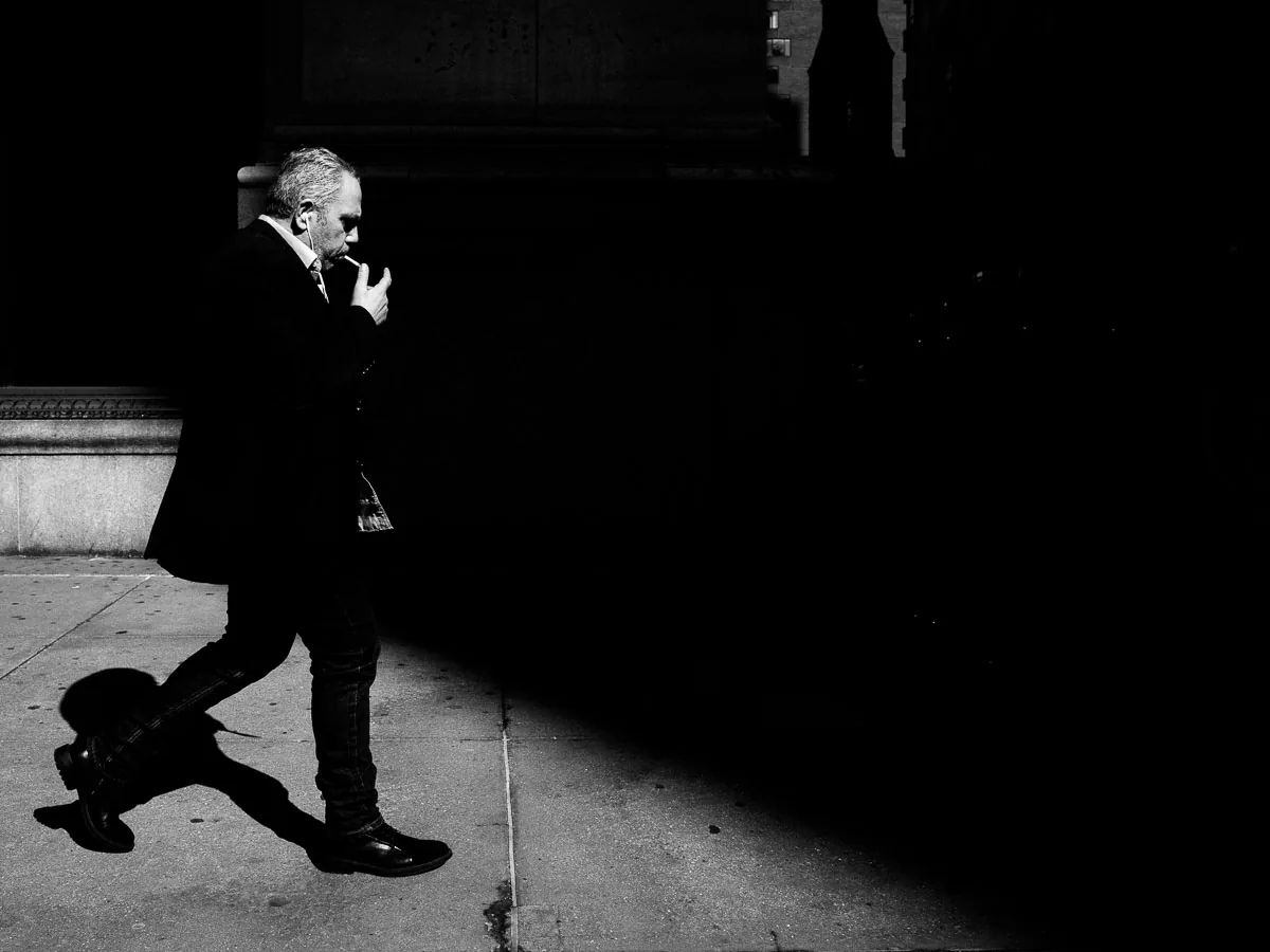 Black-and-white NYC street photograph of a smoking man crossing into deep shadow