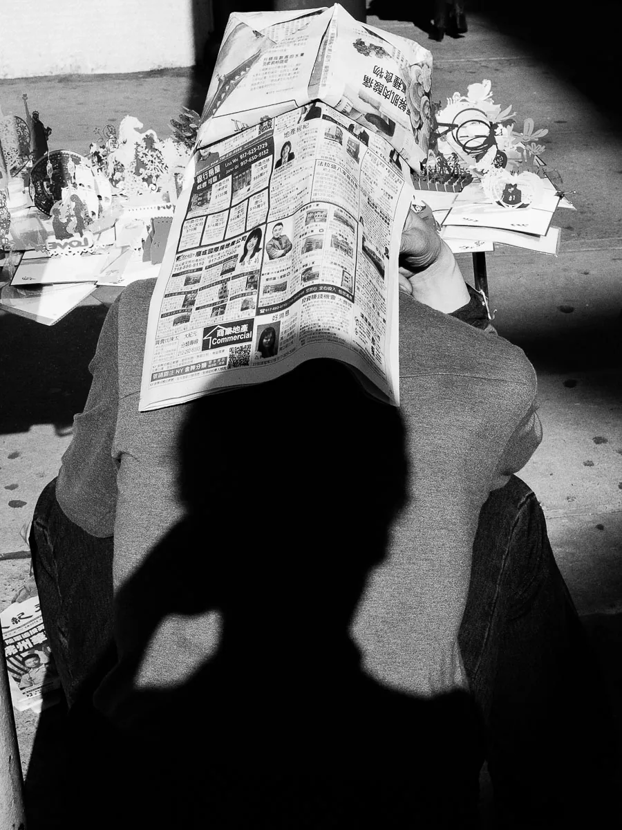 Black-and-white Chinatown street photograph of a newspaper reader covered by a shadow