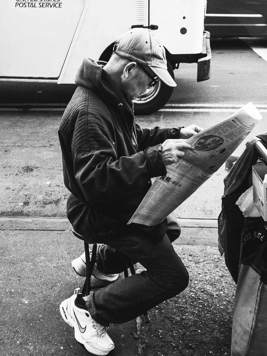 Black-and-white NYC street portrait of a man reading a newspaper beside the road