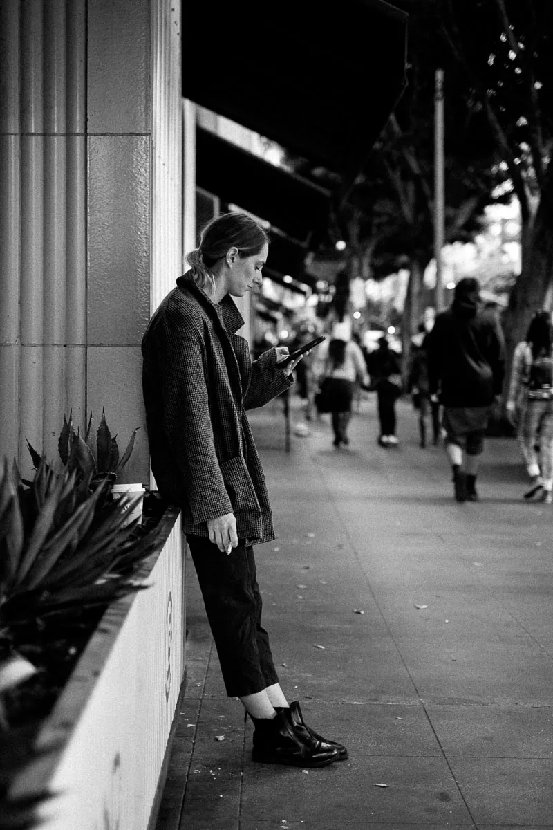 Candid NYC street photograph of a woman checking her phone beneath an elevated railway